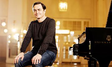 Pierre-Laurent Aimard sat next to a piano, facing the camera and smiling with his hands on his knees