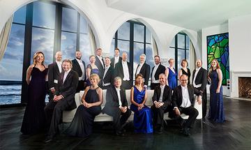 Multiple members of The Sixteen; some are sat down on a white sofa, and most are stood at the back in front of large windows