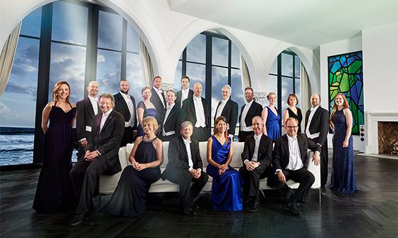 Multiple members of The Sixteen; some are sat down on a white sofa, and most are stood at the back in front of large windows