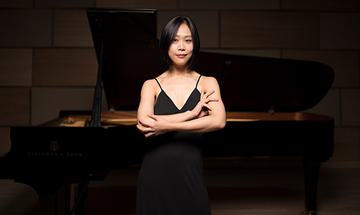 Yeol Eum Son stood in front of a piano with her arms crossed, wearing a black outfit and smiling at the camera