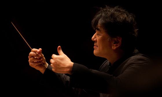 Yutaka Sado with his eyes closed and both hands raised as one is holding a baton, on a dark background