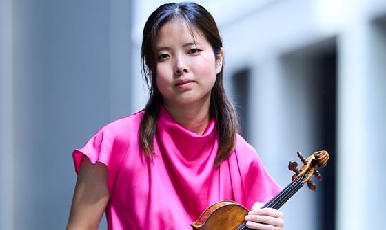 Hana Chang, a woman in a vibrant pink top holding a violin, against a blurred, light-coloured background