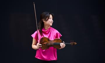 Hana Chang, a woman in a bright pink blouse holding a violin and bow, standing against a dark background. She gazes to the side with a smiling expression
