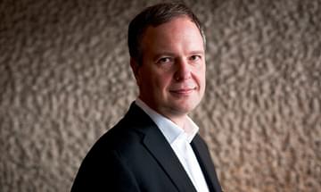 Sakari Oramo in a suit standing against a textured neutral wall, looking at the camera and smiling softly