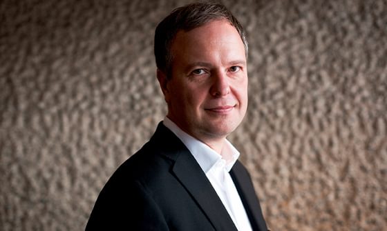 Sakari Oramo in a suit standing against a textured neutral wall, looking at the camera and smiling softly