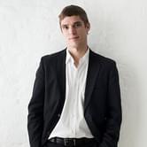 Cédric Tiberghien wearing a black suit with an open-collared white shirt, leaning against a white textured wall. He is smiling softly at the camera
