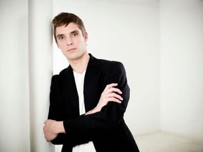 Cédric Tiberghien with short dark hair leaning against a white pillar in a minimalistic white room. He is wearing a black blazer over a white shirt, arms casually crossed