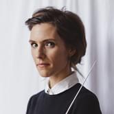 Chloé Van Soeterstède, a woman with short brown hair, wearing a black jumper over a white collared shirt, holding a conductor's baton. She is standing against a light curtain backdrop