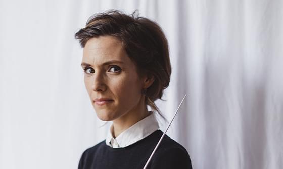 Chloé Van Soeterstède, a woman with short brown hair, wearing a black jumper over a white collared shirt, holding a conductor's baton. She is standing against a light curtain backdrop