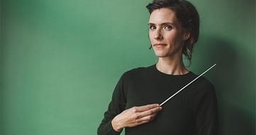 Conductor Chloé Van Soeterstède wearing a dark green jumper, holding a baton and standing against a green background, smiling softly at the camera