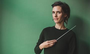 Conductor Chloé Van Soeterstède wearing a dark green jumper, holding a baton and standing against a green background, smiling softly at the camera