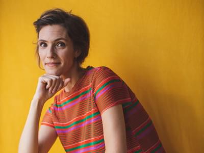Chloé Van Soeterstède wearing a vibrant striped top and yellow trousers, posing against a matching yellow backdrop and smiling at the camera with her chin resting on her hand