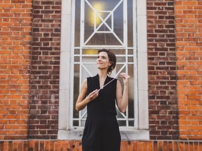 Chloé Van Soeterstède wearing a black sleeveless outfit, standing against a red brick wall with an ornate window, holding a baton and gazing upward