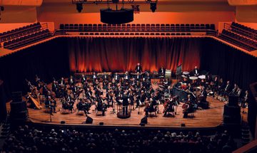 The BBC Concert Orchestra performing on a warmly lit stage with musicians seated in semi-circles, and an audience watching