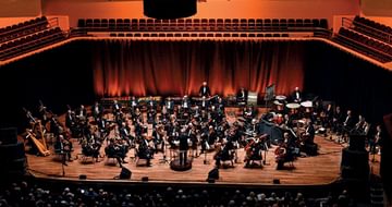 The BBC Concert Orchestra performing on a warmly lit stage with musicians seated in semi-circles, and an audience watching