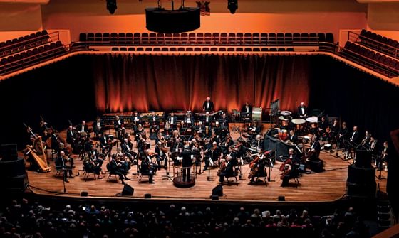 The BBC Concert Orchestra performing on a warmly lit stage with musicians seated in semi-circles, and an audience watching