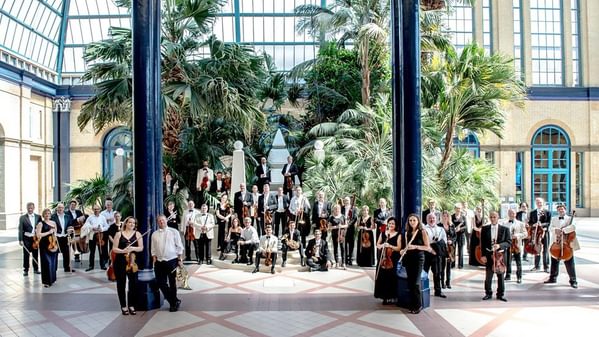 The BBC Concert Orchestra members posing in a sunlit conservatory with tall palm trees behind them. The musicians are holding instruments like violins and cellos, in an informal setting