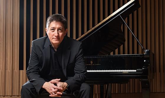 Freddy Kempf wearing a black suit sitting on a piano bench, hands clasped, in front of a grand piano and a wooden panel backdrop