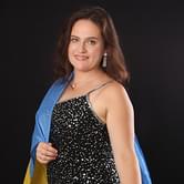 Maria Pukhlyanko, a woman in a sparkly black dress with a Ukrainian flag draped around her shoulders, looking at the camera and smiling, against a dark background