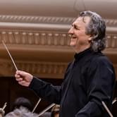 Volodymyr Sirenko in a black suit holding a baton, leading an orchestra and smiling