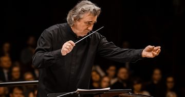 Volodymyr Sirenko with grey hair conducting an orchestra, holding a baton. He's wearing a black shirt