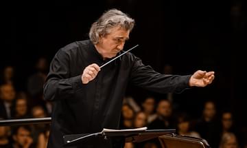 Volodymyr Sirenko with grey hair conducting an orchestra, holding a baton. He's wearing a black shirt