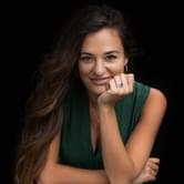 Nicola Benedetti, a woman in a green dress, with long brown hair, leaning on a wooden block. She is resting her chin on her hand, wearing a blue ring, against a black background