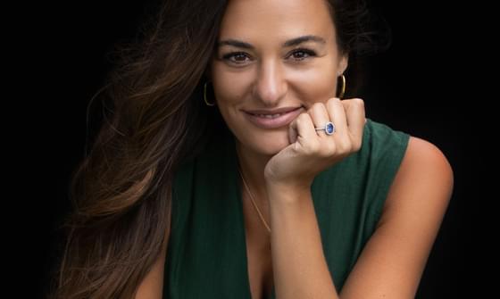 Nicola Benedetti, a woman in a green dress, with long brown hair, leaning on a wooden block. She is resting her chin on her hand, wearing a blue ring, against a black background