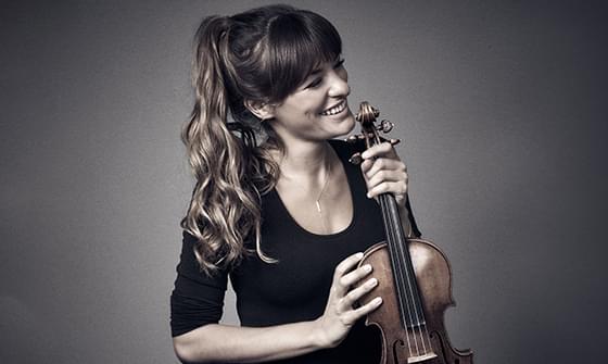 Nicola Benedetti, a woman with long dark hair in a ponytail, smiling and holding a violin. She is wearing a black top and smiling away from the camera