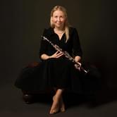 Katherine Spencer, a woman sitting on a dark bench, holding a clarinet in hands. She is wearing a black dress and smiling gently, on a dark background