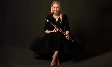 Katherine Spencer, a woman sitting on a dark bench, holding a clarinet in hands. She is wearing a black dress and smiling gently, on a dark background