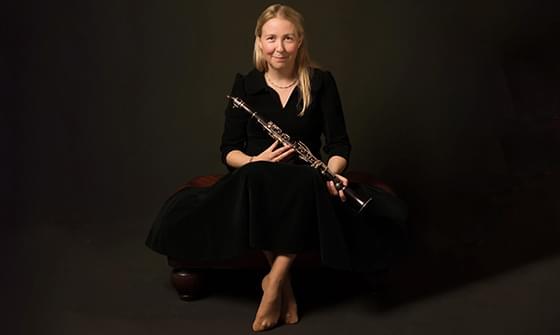 Katherine Spencer, a woman sitting on a dark bench, holding a clarinet in hands. She is wearing a black dress and smiling gently, on a dark background
