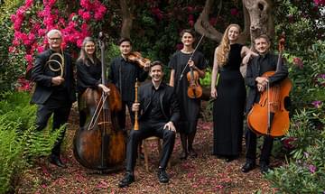 A group of seven musicians in black attire standing and sitting among lush greenery and vibrant pink flowers, holding classical instruments