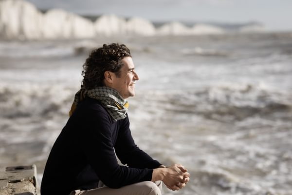 Robin Ticciati wearing a scarf and dark jumper, sitting and smiling by a choppy sea