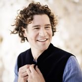 Robin Ticciati with dark curly hair wearing a blue shirt and dark jacket. He is smiling softly away from the camera, on a neutral blurred background
