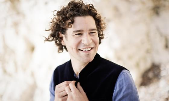 Robin Ticciati with dark curly hair wearing a blue shirt and dark jacket. He is smiling softly away from the camera, on a neutral blurred background