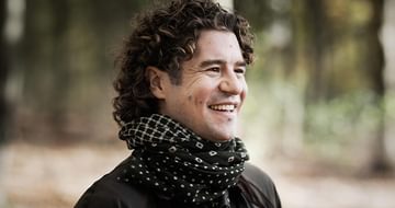 Robin Ticciati, a man with dark curly hair, wearing a patterned scarf and dark jacket, standing outdoors in a blurred forest. He is smiling away from the camera