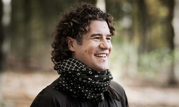 Robin Ticciati, a man with dark curly hair, wearing a patterned scarf and dark jacket, standing outdoors in a blurred forest. He is smiling away from the camera