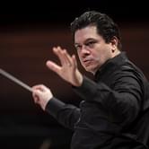 Conductor Cristian Măcelaru in a black shirt passionately leading an orchestra, baton in hand, on a dark background