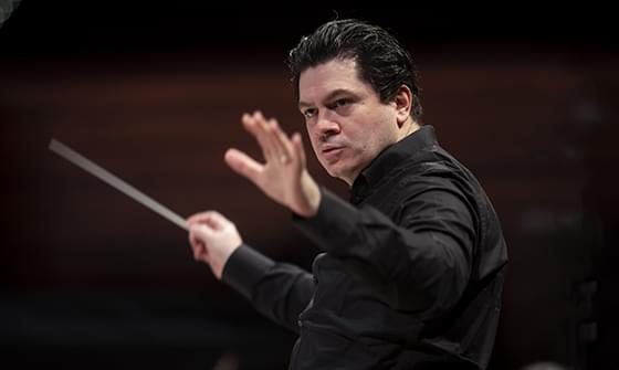 Conductor Cristian Măcelaru in a black shirt passionately leading an orchestra, baton in hand, on a dark background