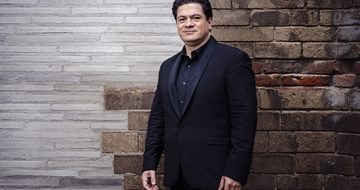Cristian Măcelaru in a dark suit standing against a textured brick wall, looking towards the camera with a soft smile