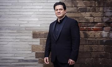 Cristian Măcelaru in a dark suit standing against a textured brick wall, looking towards the camera with a soft smile