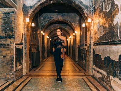 Nicola Benedetti in a black, asymmetric dress standing in an arched, dimly lit hallway, holding a violin