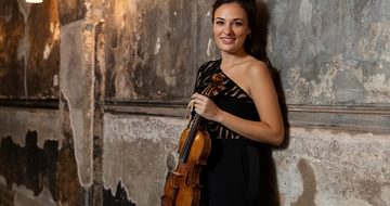 Nicola Benedetti wearing an elegant black dress and smiling at the camera, holding a violin in a dimly lit corridor with rustic, textured walls and soft, warm lighting from overhead lamps