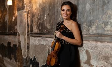 Nicola Benedetti wearing an elegant black dress and smiling at the camera, holding a violin in a dimly lit corridor with rustic, textured walls and soft, warm lighting from overhead lamps