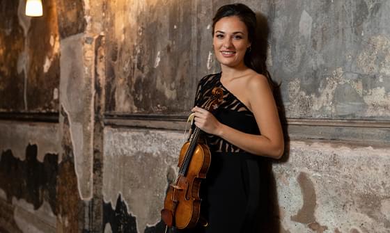 Nicola Benedetti wearing an elegant black dress and smiling at the camera, holding a violin in a dimly lit corridor with rustic, textured walls and soft, warm lighting from overhead lamps