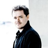 Benjamin Grosvenor, a man with short, curly dark hair and a subtle smile, wearing a dark jacket, standing against a blurred light background