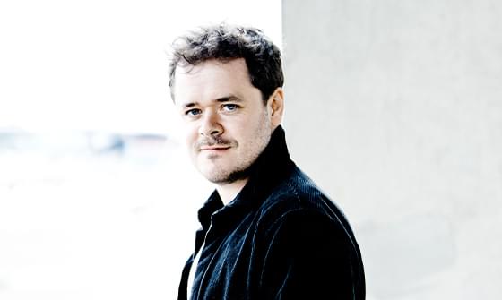 Benjamin Grosvenor, a man with short, curly dark hair and a subtle smile, wearing a dark jacket, standing against a blurred light background