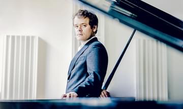 Benjamin Grosvenor in a suit standing pensively behind a grand piano, lid open, on a bright neutral background