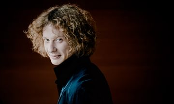 Santtu-Matias Rouvali with brown curly hair, wearing a dark jacket and smiling subtly over his shoulder, on a dark background